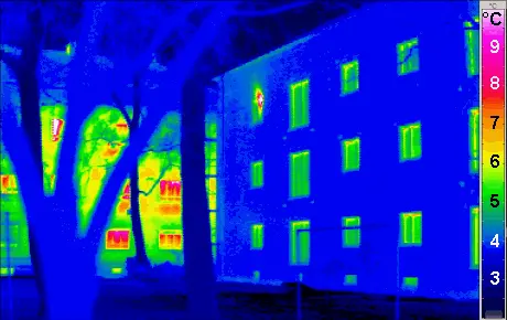 A thermal-camera image of two buildings on a cold day, with a temperature scale from about 3 to 9 degrees Celsius. The well-insulated building on the right is mostly dark blue, with only its windows slightly warmer. The older building on the left, seen behind bare trees, glows yellow and red, especially around its windows.