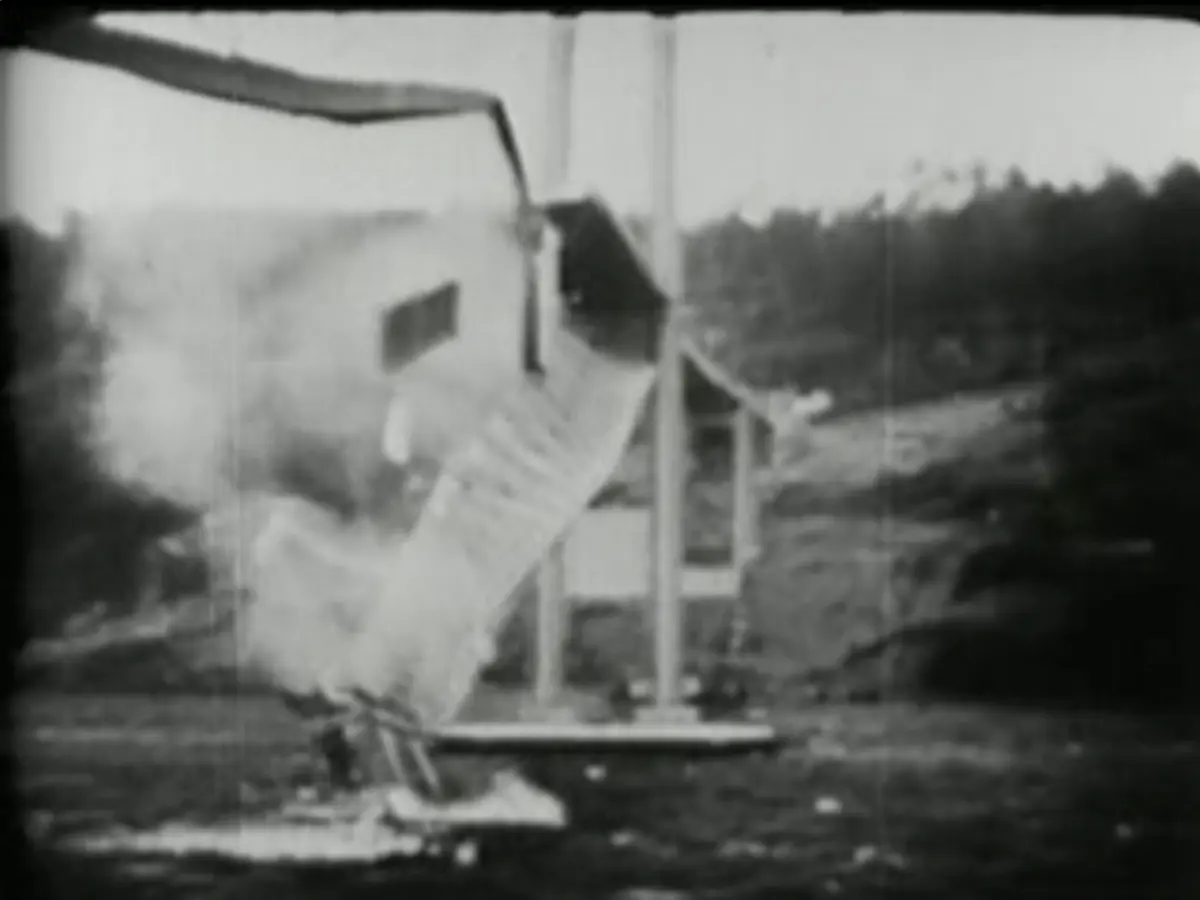 A black-and-white film still of the Tacoma Narrows Bridge collapsing in 1940. A long section of the road deck is twisting and falling between the towers, with a cloud of debris below it.