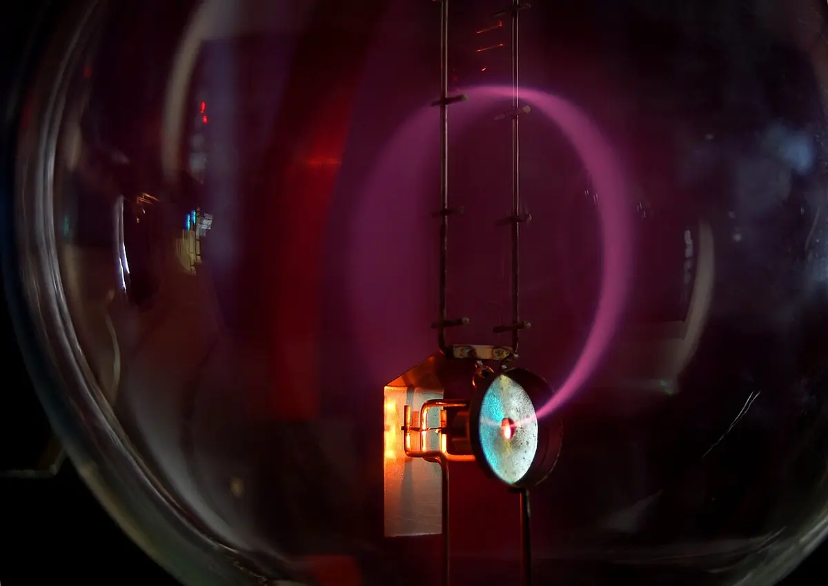 A glass bulb in a darkened room. An electron gun near the bottom fires a beam that glows violet-pink as it travels round an almost complete circle inside the bulb. A vertical ladder of measuring rungs stands in the bulb behind the beam.
